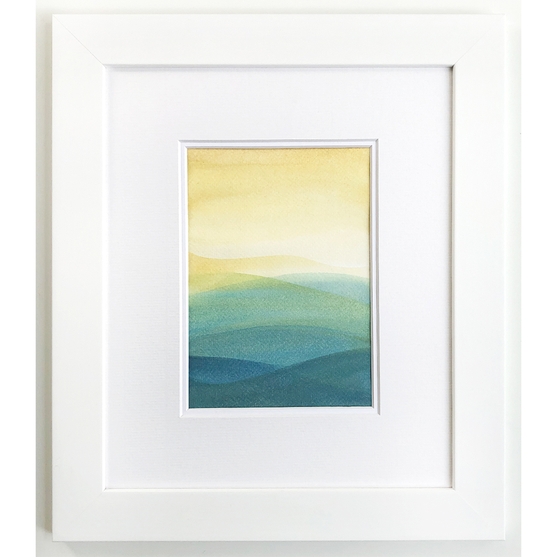 Watercolor Painting of a yellow and white sky setting over green, blue, and teal layered mountains. All framed and matted in white.